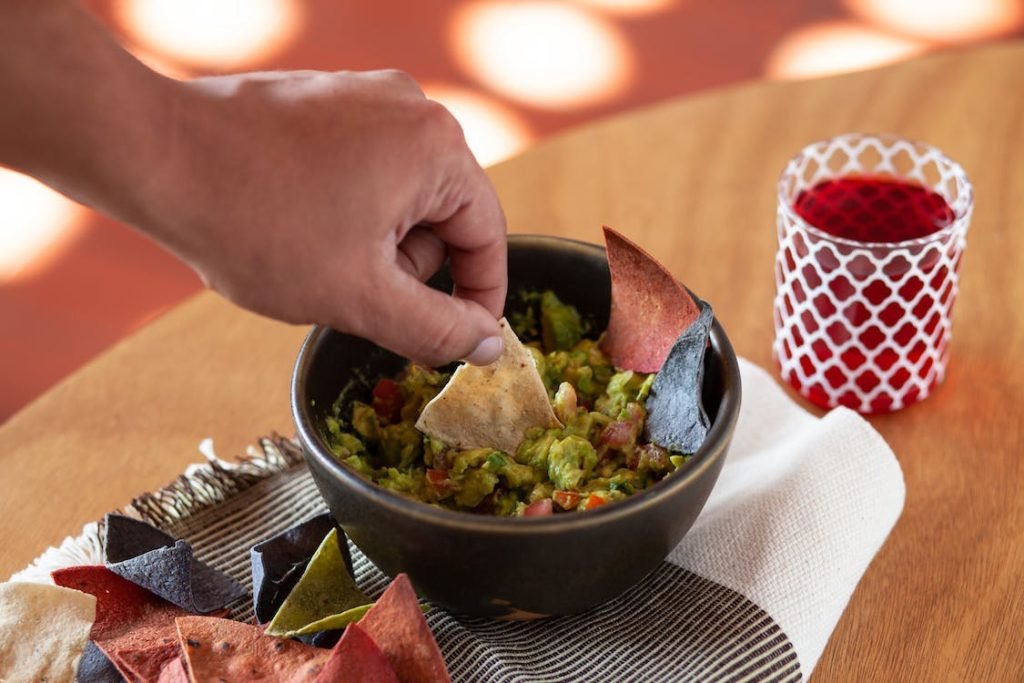 A hand dipping a triangular tortilla chip into a bowl of homemade cheesy spinach and artichoke dip, surrounded by multicolored chips, on a textured tablecloth with a refreshing beverage in the background.