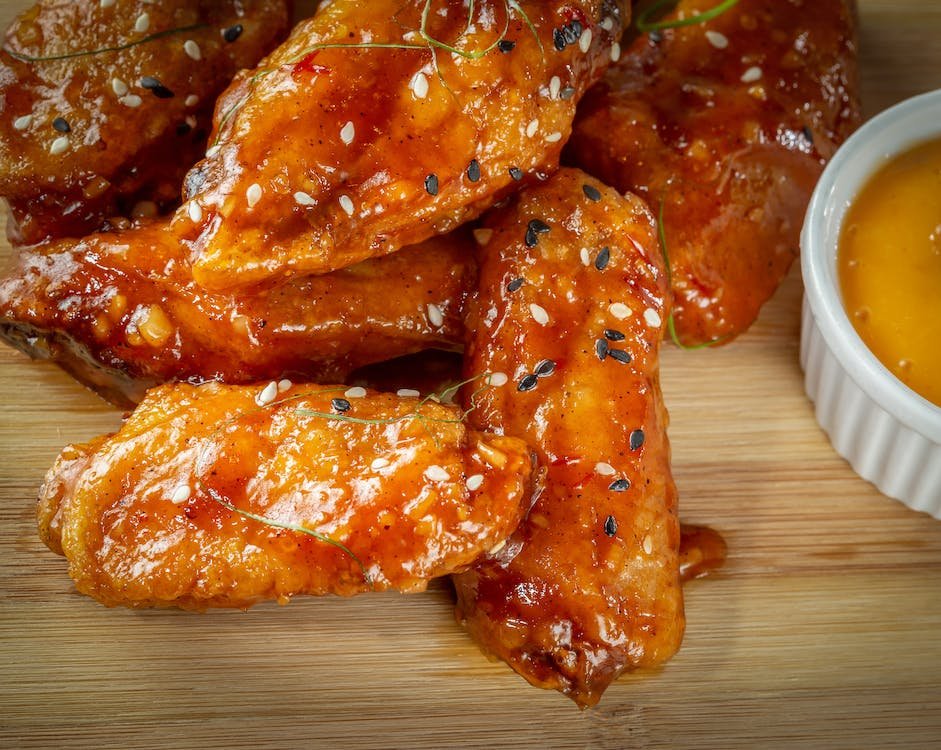 Close-up of juicy, glazed chicken wings garnished with sesame seeds and herbs, besides a dipping sauce, ideal for Happy Hour snacking.