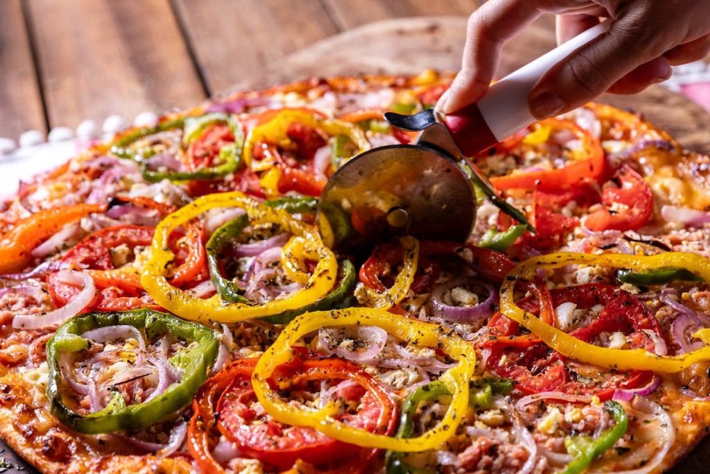 Freshly prepared Diavola Pizza, rich with colorful bell peppers, onions, and spicy sausage, being sliced on a rustic wooden table, ready to enhance any happy hour gathering.