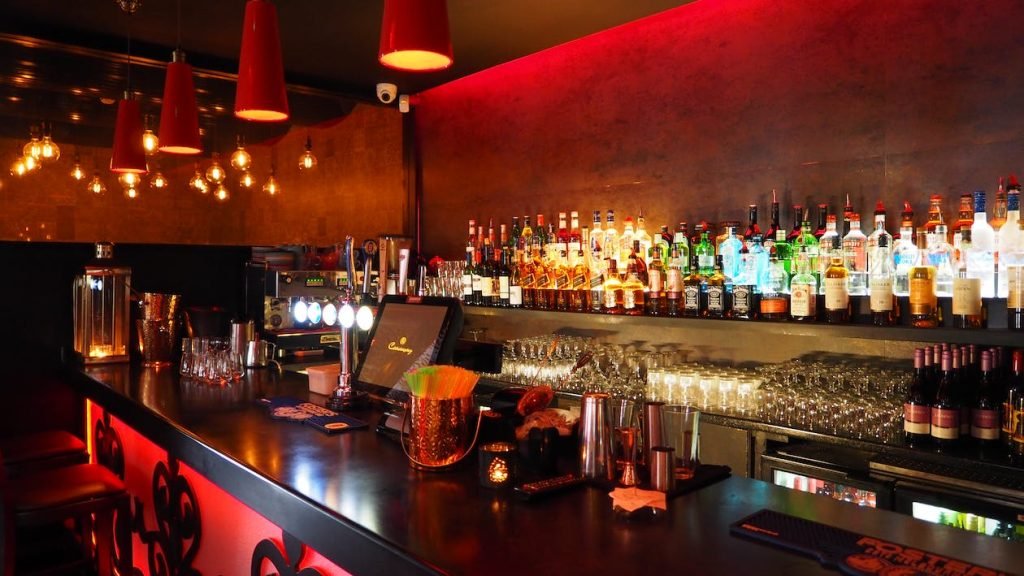 A stylish downtown bar and lounge with ambient lighting, red pendant lamps, and a well-stocked assortment of drinks creates an inviting atmosphere for social evenings.