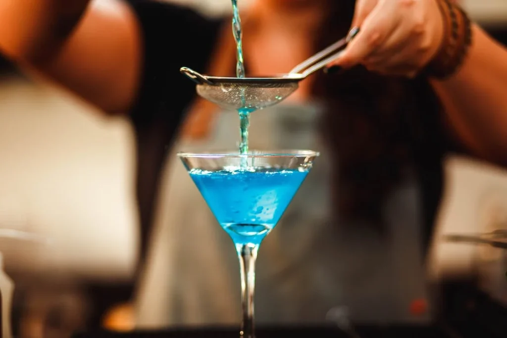 A bartender masterfully strains a bright blue cocktail into a martini glass, showcasing the precision and flair of cocktail crafting.