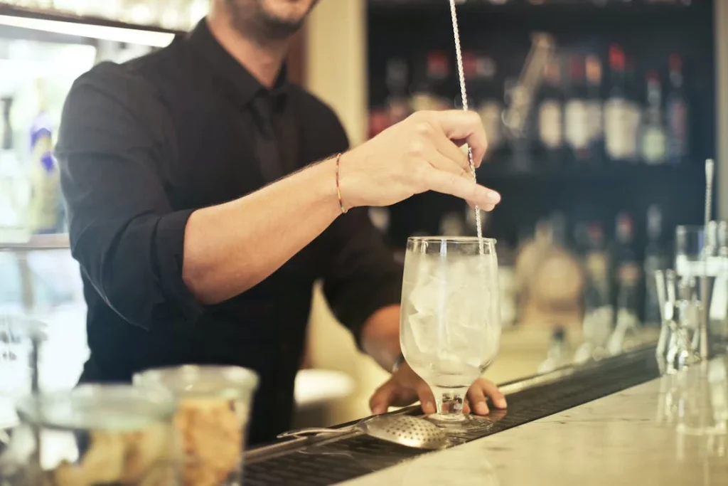 Bartender in a black shirt skillfully stirring a tall glass of ice, preparing a signature cocktail with precision against a backdrop of an assorted liquor collection.